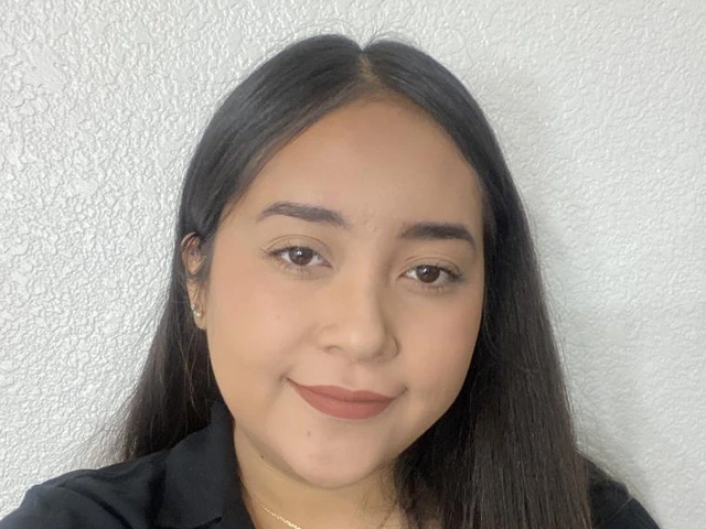 A woman with straight, long dark hair smiles softly in front of a light-textured wall. She is wearing a black blouse and a gold necklace with a letter "R" charm.