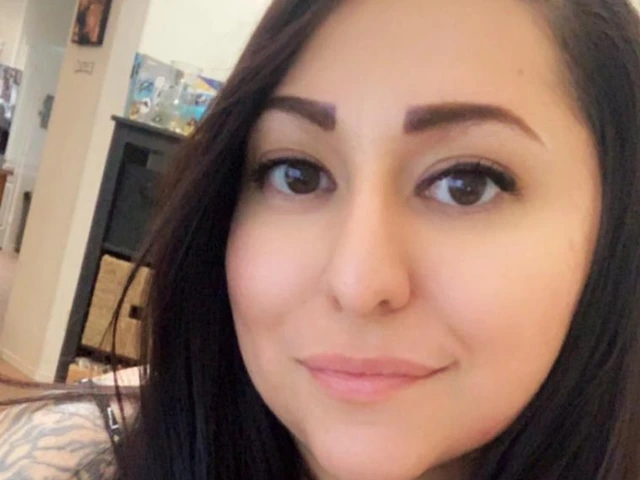 A woman with long dark hair and tattoos on her arm smiles gently while seated indoors. She is wearing a sleeveless black top and is in a warmly lit room with furniture and artwork in the background.