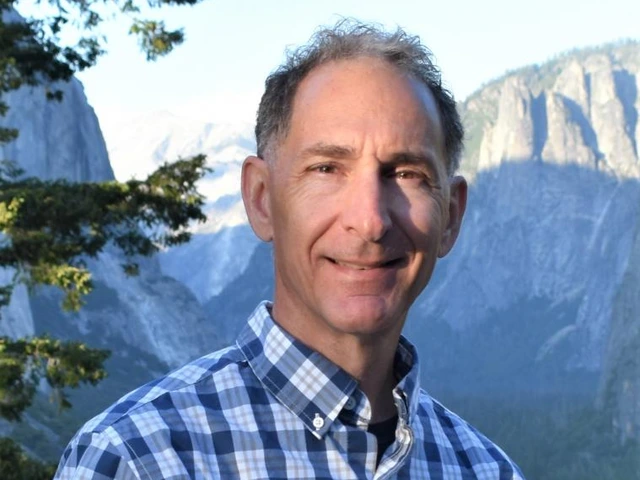Man wearing a checkered shirt smiling in front of a mountain landscape.