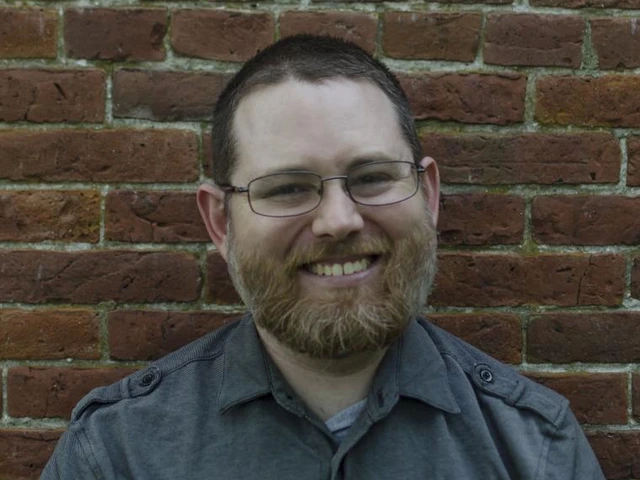 Man with glasses and a beard smiling in front of a red brick wall.