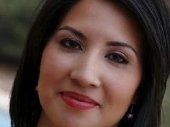 Close-up of a woman with long dark hair and pearl earrings, smiling in an outdoor setting.