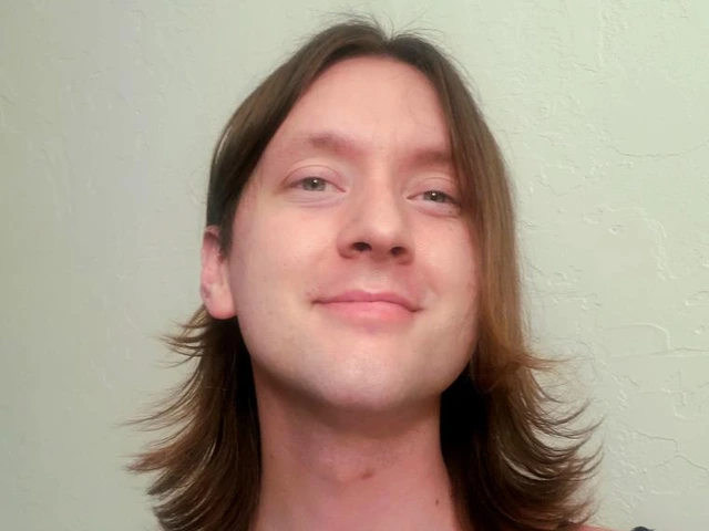 A person with shoulder-length, straight brown hair smiles slightly while standing in front of a light-colored wall. They are wearing a black and white striped top with thin black straps.