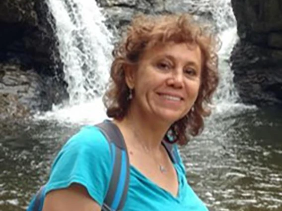 Cora Varas-Nelson smiling near a waterfall, wearing a turquoise shirt and backpack.