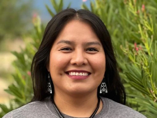 A woman with long black hair smiles while standing outdoors in front of green foliage with pink flowers. She is wearing a gray shirt and turquoise jewelry, including a pendant necklace and silver earrings.