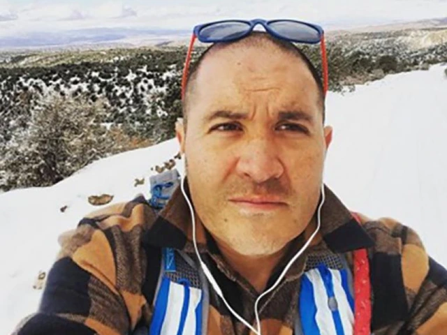 Close-up of a man wearing a plaid jacket and blue sunglasses, standing in a snowy landscape with headphones in.