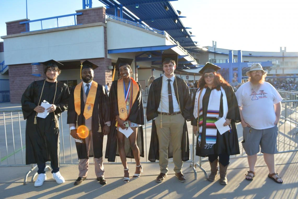 Graduates and mentors pose outdoors at Pima Community College commencement ceremony, with a stadium in the background.