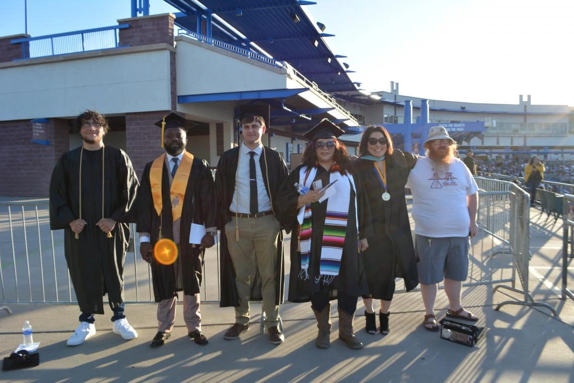 Group of graduates smiling in caps and gowns at an outdoor Pima Community College graduation ceremony.