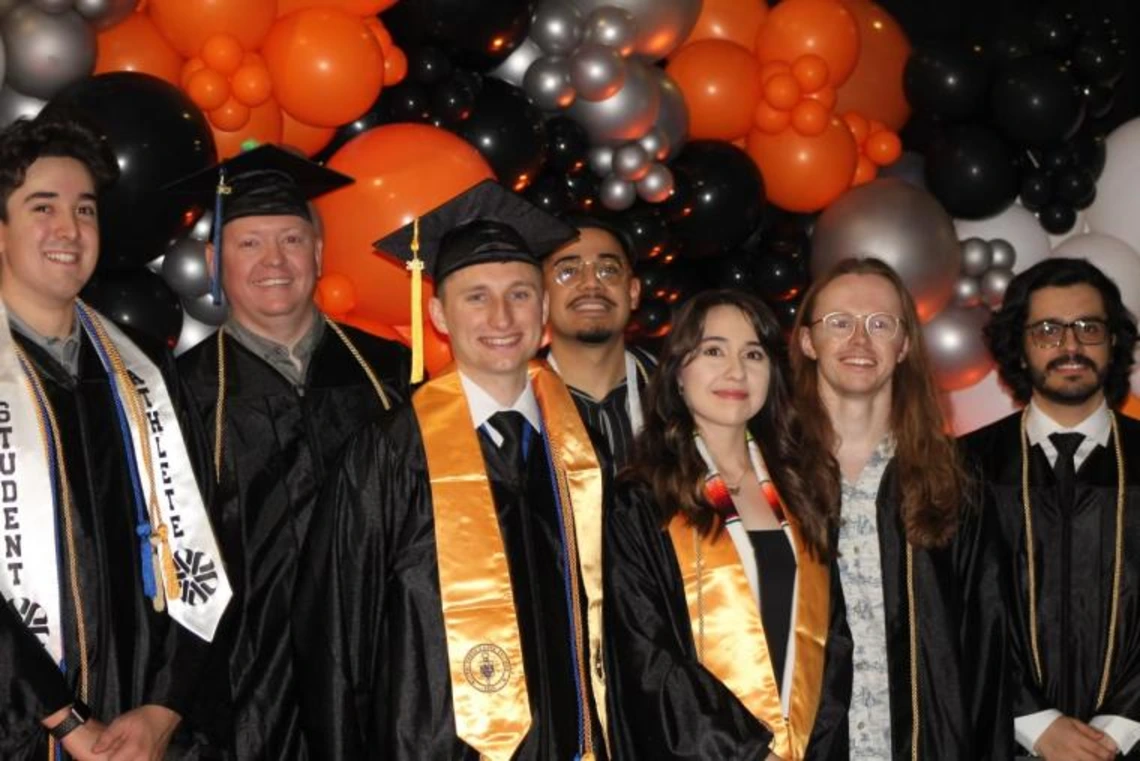 Group of graduates in caps and gowns posing in front of a black, orange, and silver balloon arch indoors.