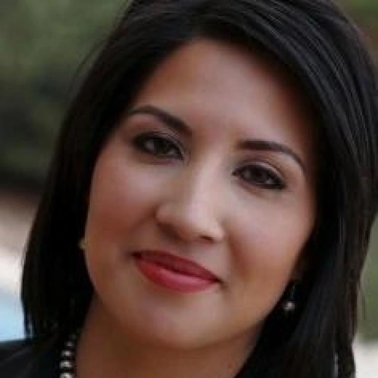 Close-up of a woman with long dark hair and pearl earrings, smiling in an outdoor setting.