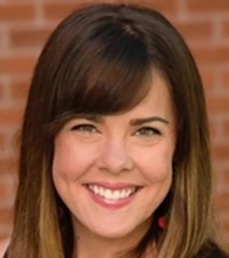 Portrait of Rachel Leih smiling in front of a brick wall, wearing a black top with bangs and shoulder-length brown hair.