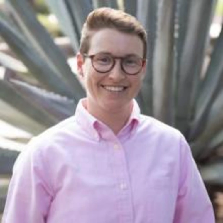 Portrait of Terrace Ewinghill in a light pink button-up shirt, smiling in front of a large agave plant.