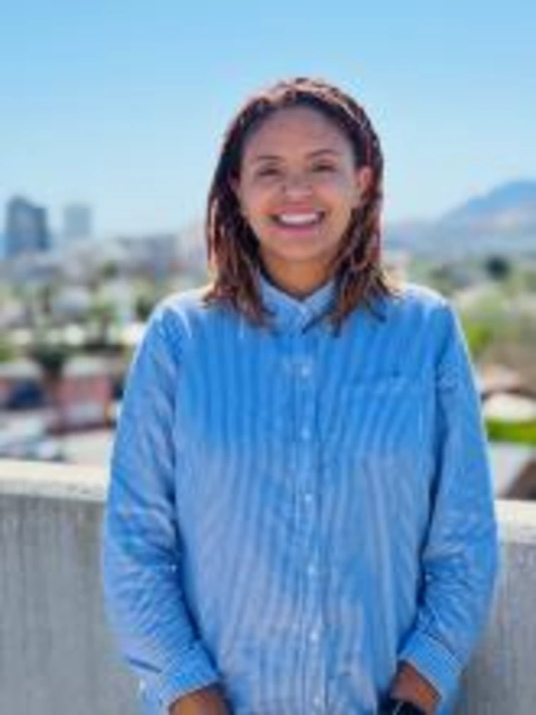 Outdoor portrait of Brenda Wadley smiling in a blue button-up shirt, with locs and a cityscape in the background.