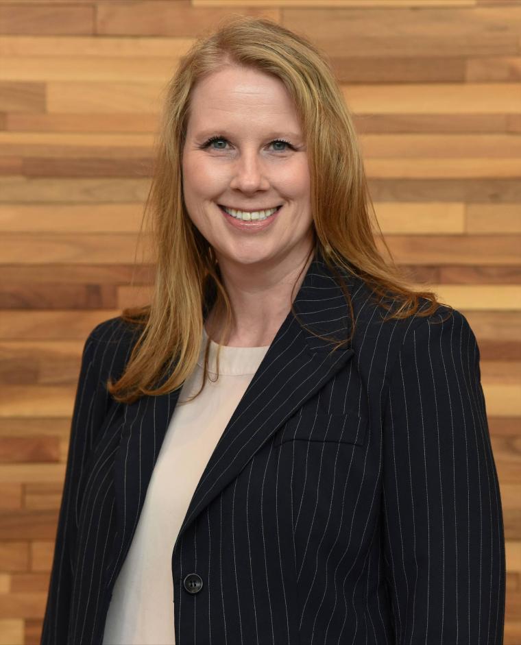 emily otts headshot, wearing a black blazer, medium blonde hair, woodgrain background